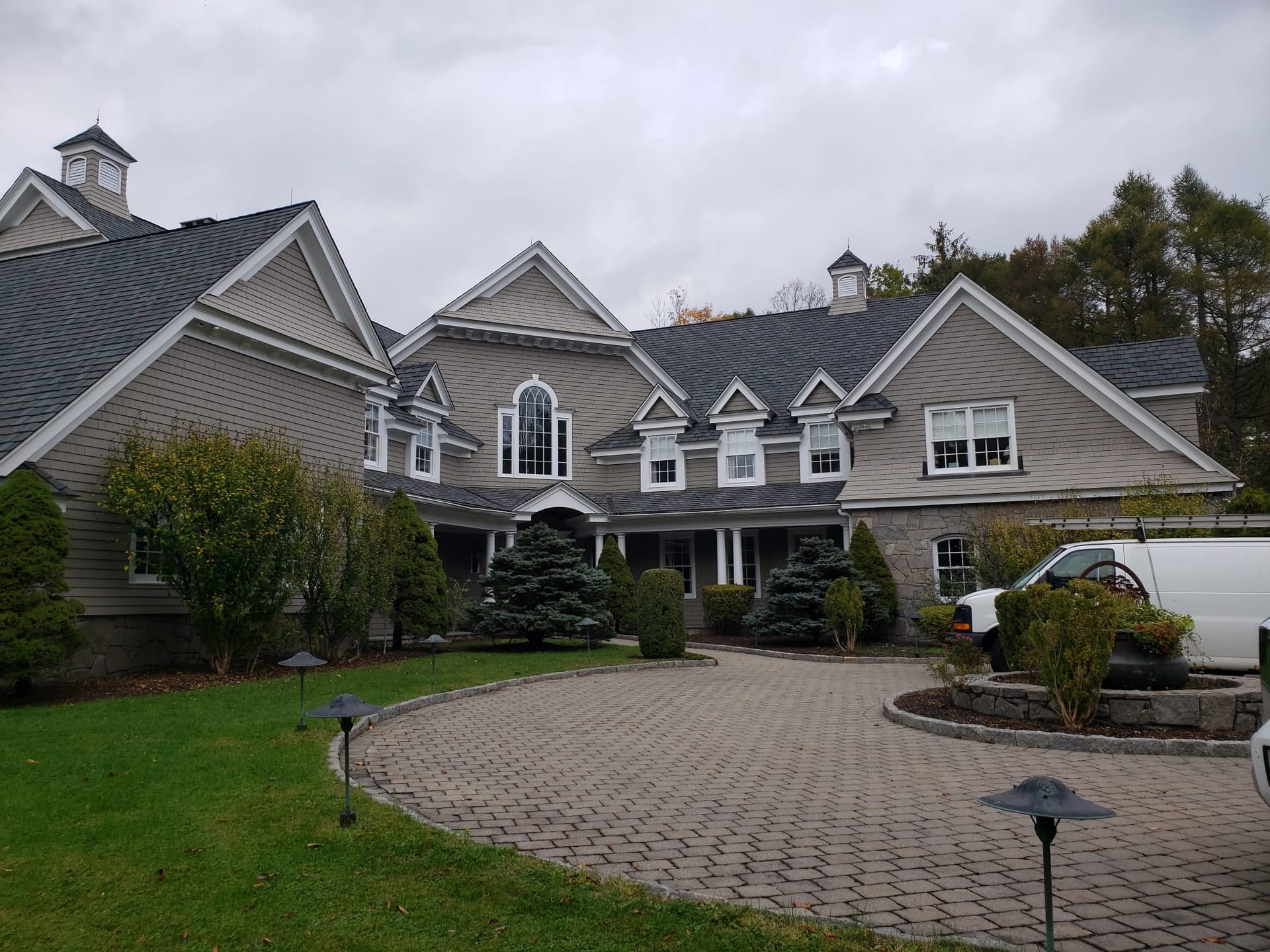 Aerial view of a two-story colonial home with newly installed gray asphalt shingle roof, brick chimney, light blue vinyl siding, white-railed rear deck with outdoor furniture, and paved driveway surrounded by mature trees